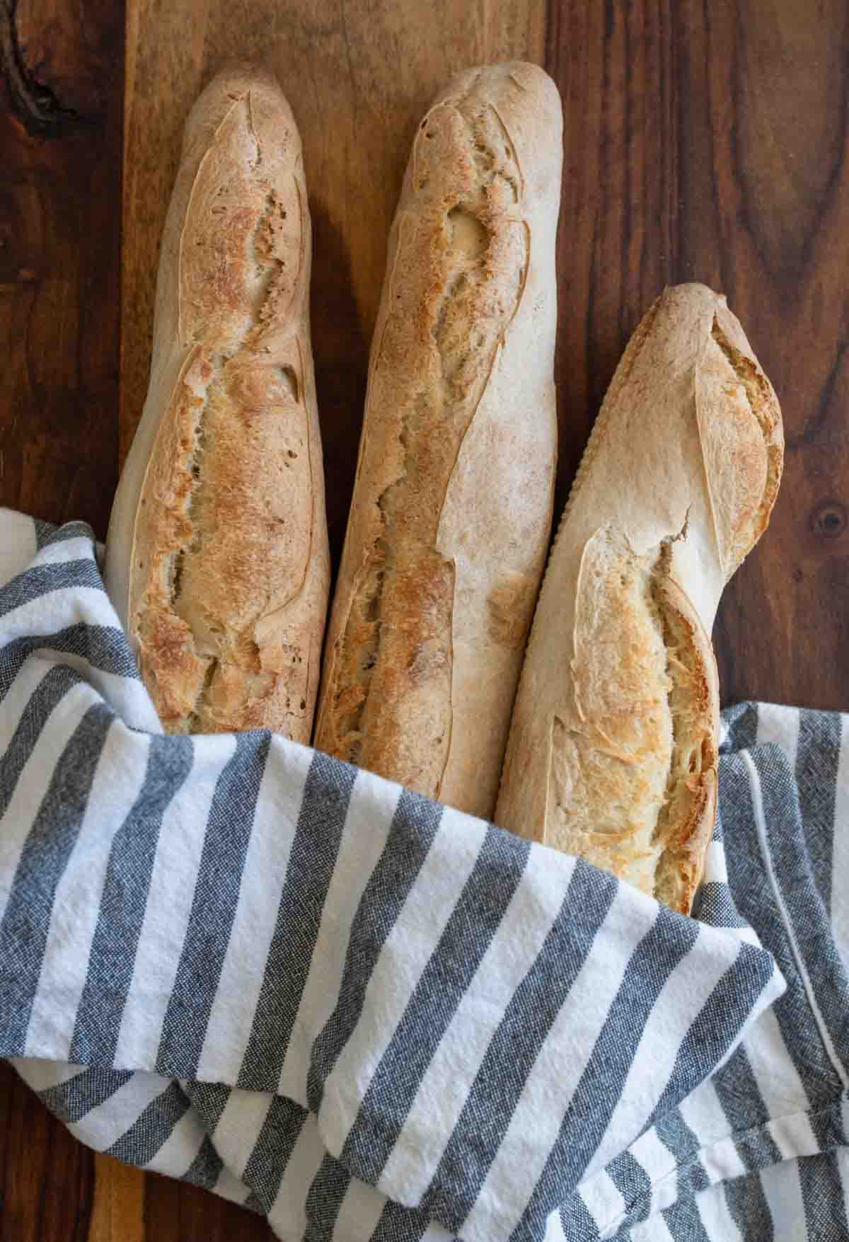 three baked baguettes in gray and white striped towel on brown countertop.