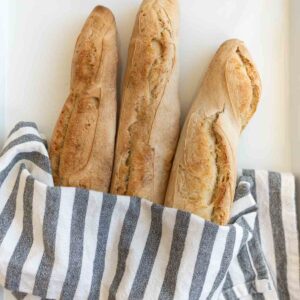 three baked baguettes in gray and white striped towel on white countertop.