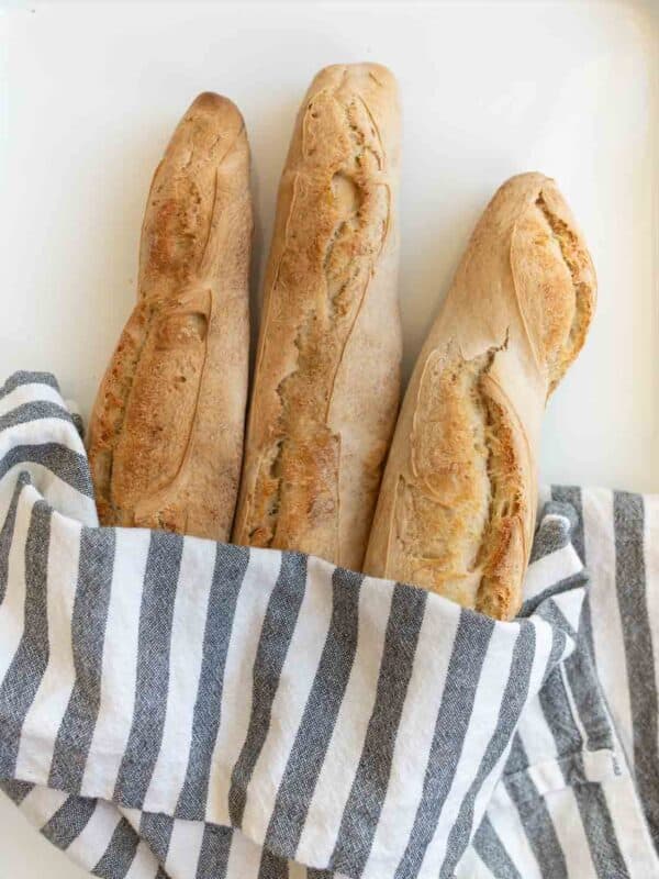 three baked baguettes in gray and white striped towel on white countertop.