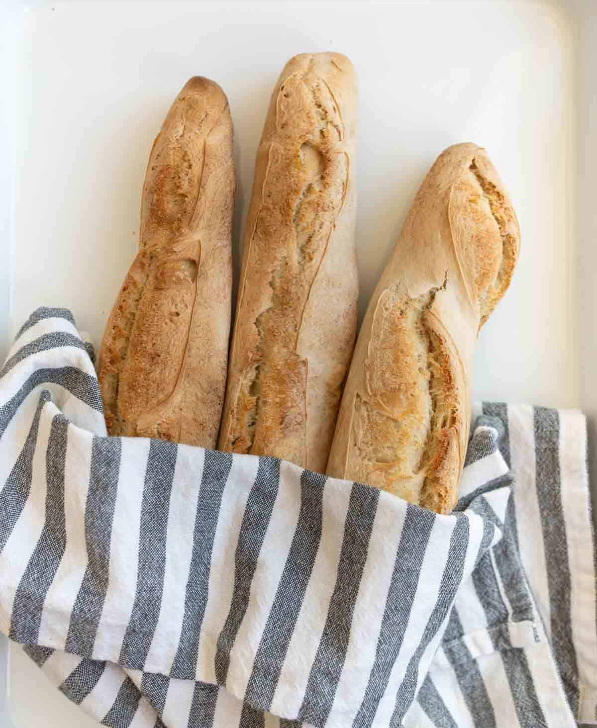 three baked baguettes in gray and white striped towel on white countertop.