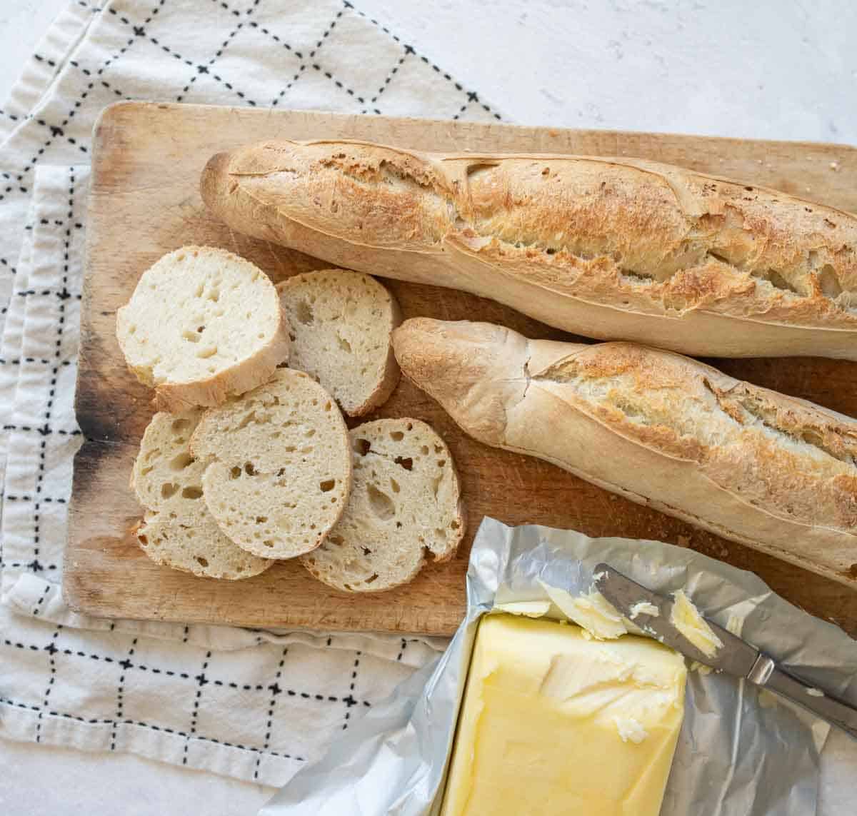 loaves of sourdough baguette with slices on brown board with butter beside.