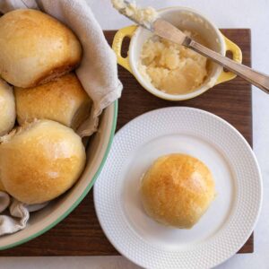 golden sourdough roll on white plate with small yellow dish with butter and a knife and bowl of rolls in the background.