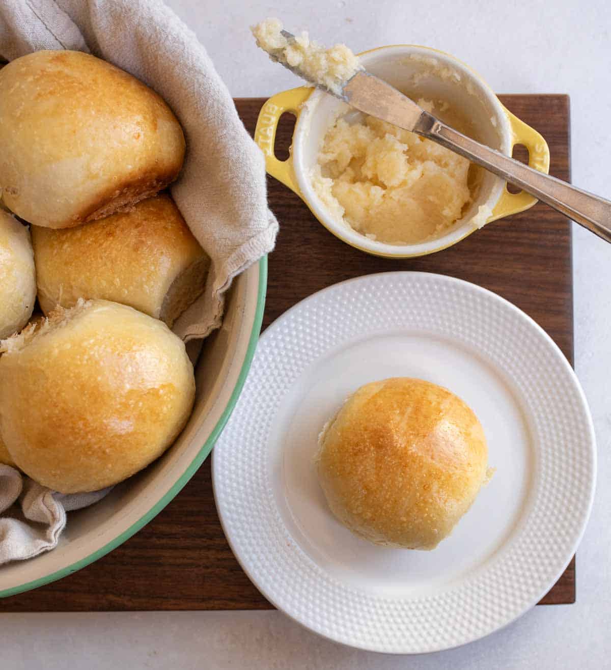 golden sourdough roll on white plate with small yellow dish with butter and a knife and bowl of rolls in the background.