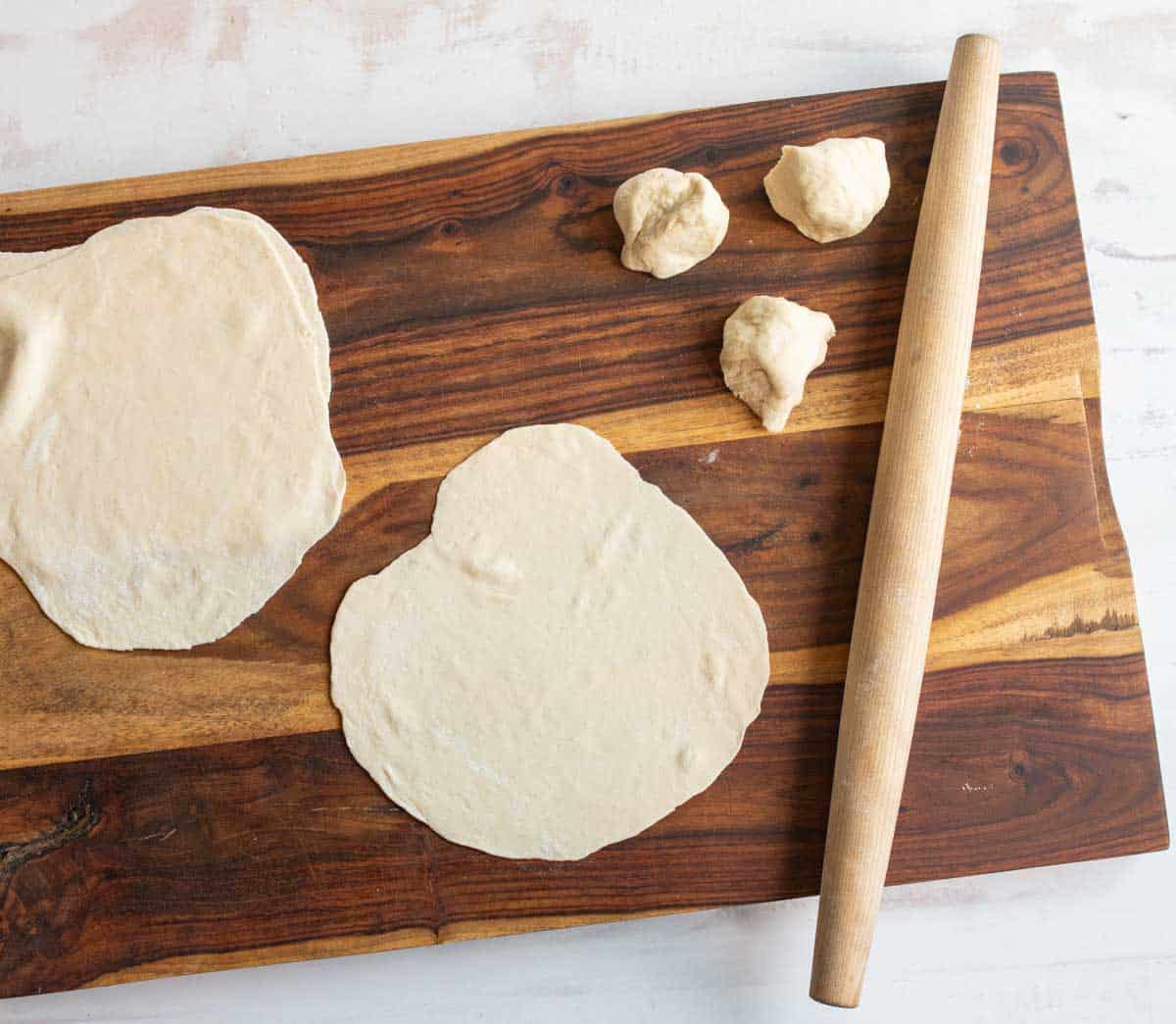 tortilla dough being rolled out with rolling pin on dark brown board.