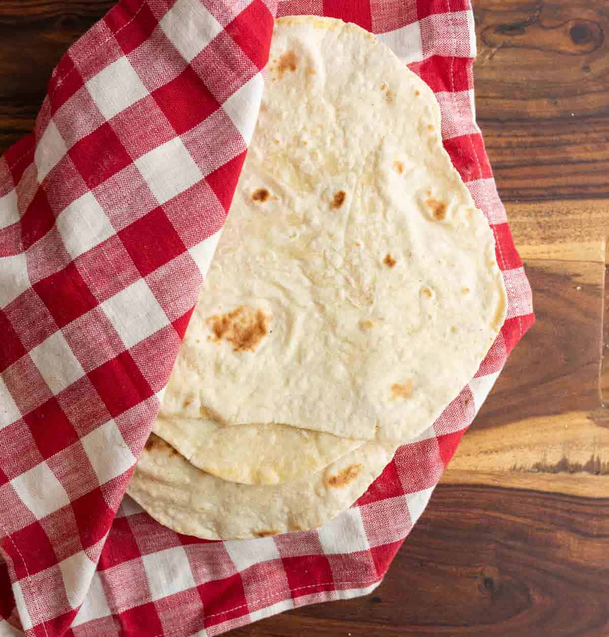 homemade sourdough tortillas being kept warm in red and white checked cloth on brown countertop.