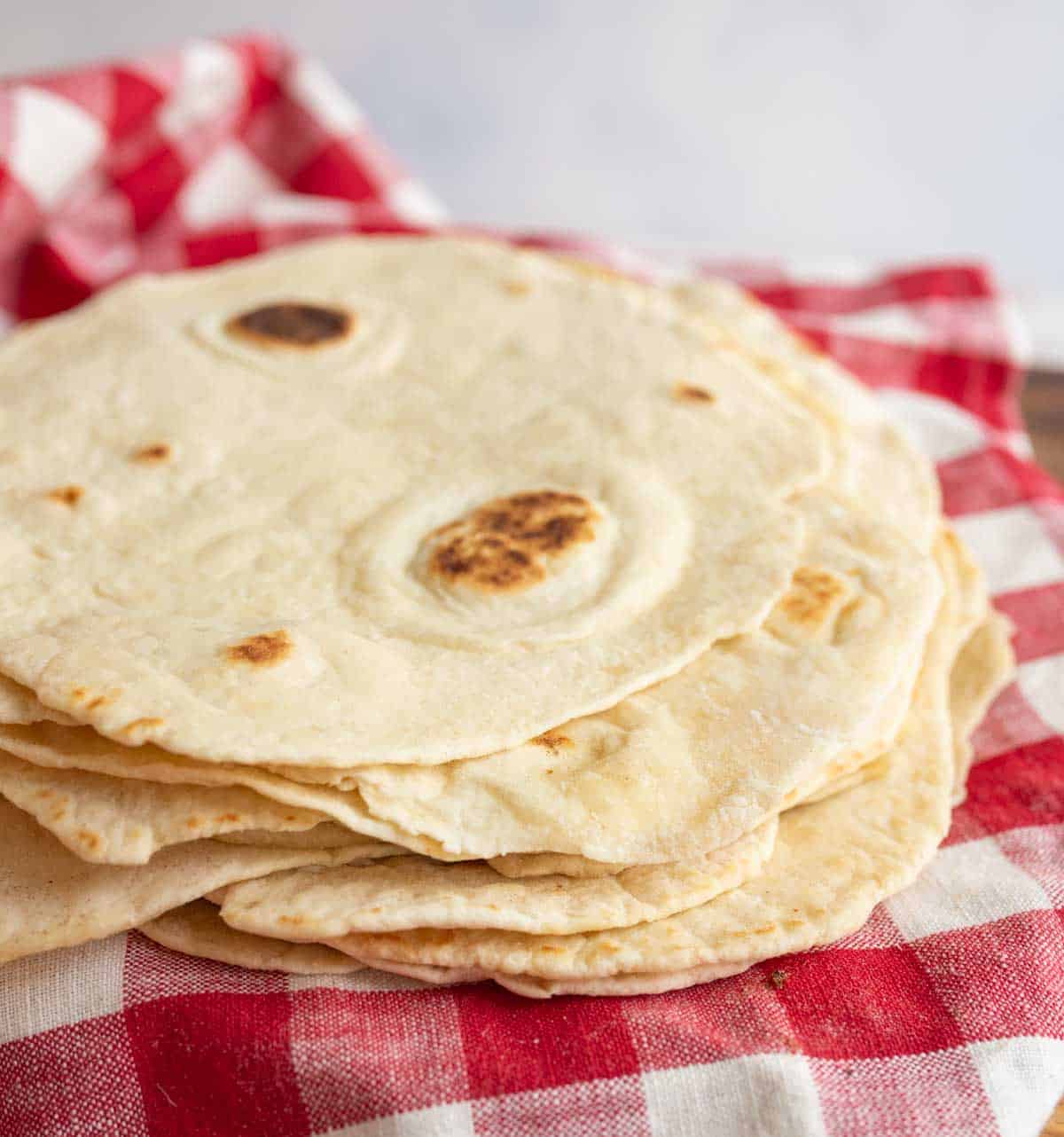 pile of cooked tortillas on red and white checked towel.