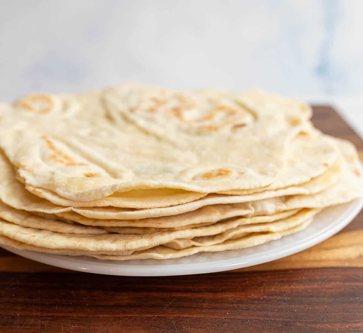 pile of tortillas on white plate sitting on brown countertop.