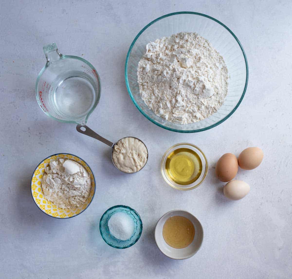 ingredients for sourdough challah bread in clear and colored bowls on white counterop.