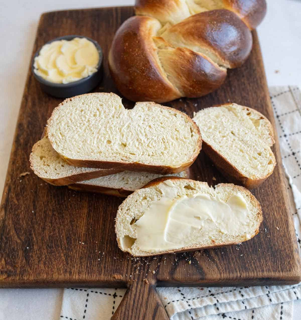 baked sourdough challah bread on brown board with slices cut off and bowl of butter.