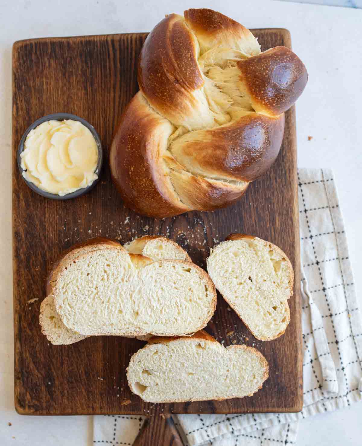 baked sourdough challah bread on brown board with slices cut off and bowl of butter.