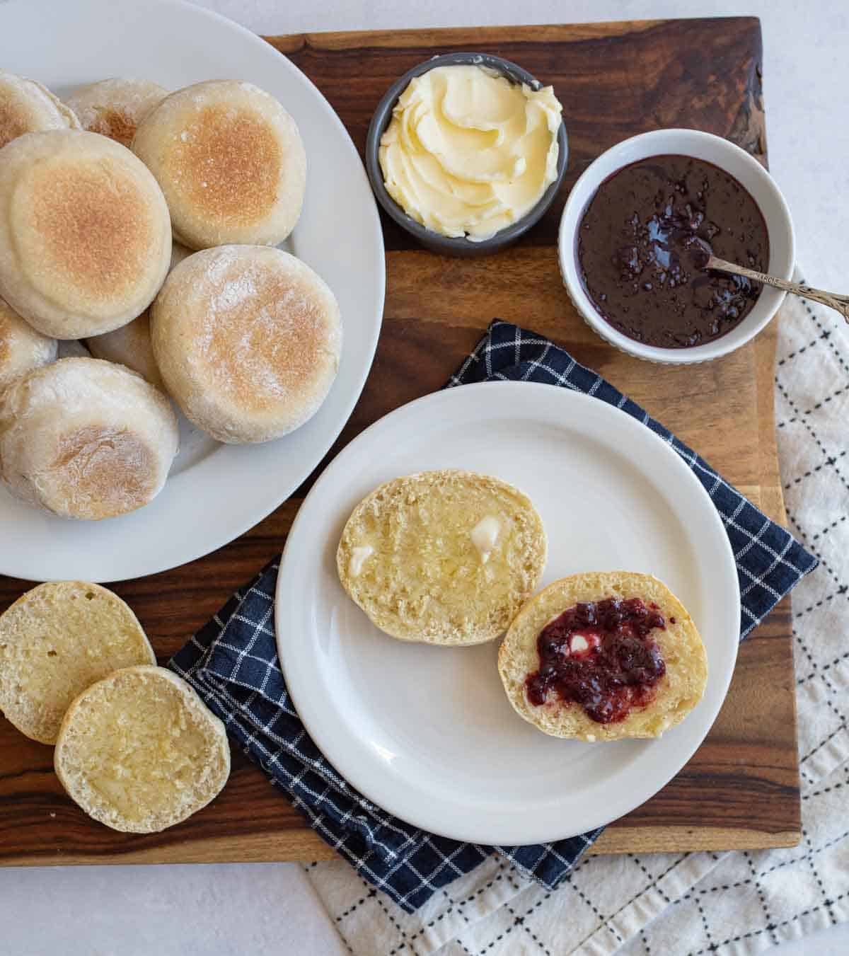 split baked english muffin on white plate with butter and jelly and pile of muffins on white plate, butter and jelly in background.