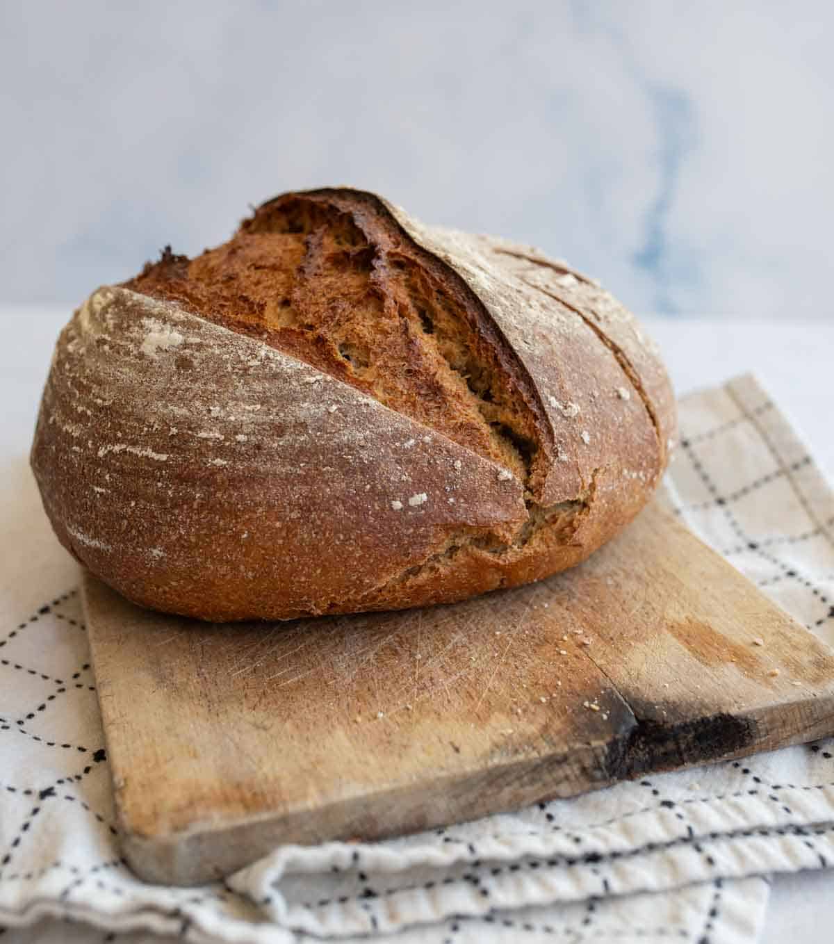 round baked loaf of rye bread on wooden cutting board set on white towel.