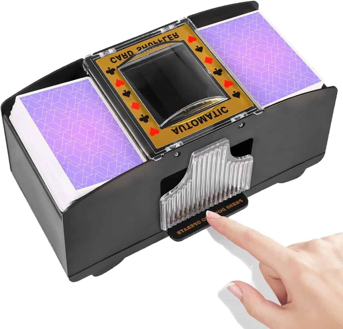 A hand presses the button on an automatic card shuffler containing two decks of playing cards with purple backs. The shuffler is black with a transparent card holder and yellow label featuring card suits.