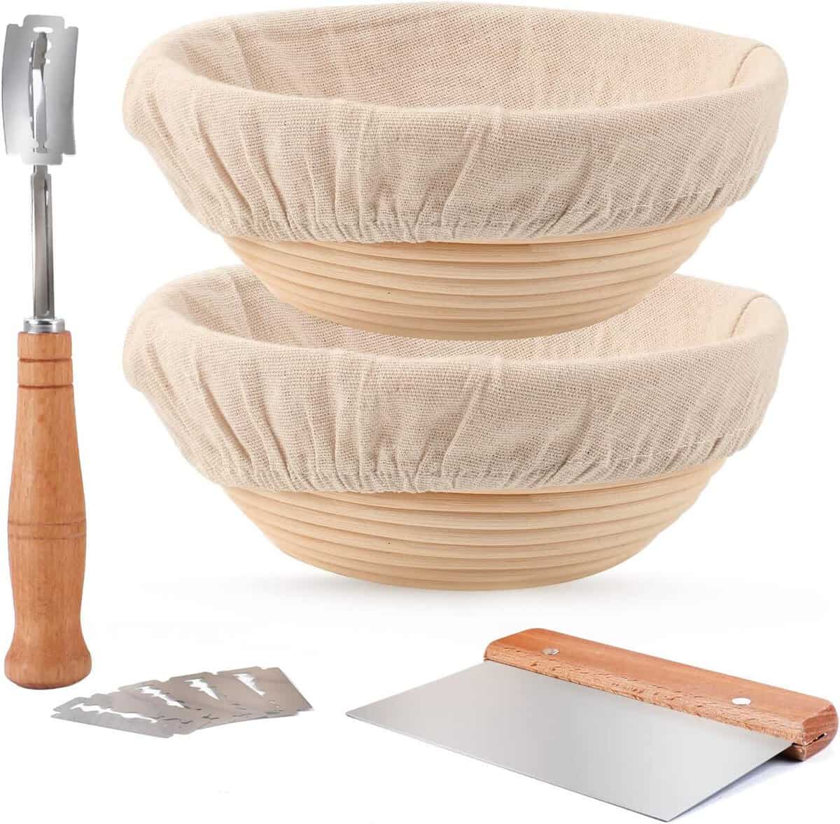 Two banneton proofing baskets with cloth liners stacked together, a metal dough scraper with a wooden handle, and a bread lame with a wooden handle. Four replacement blades are also visible.