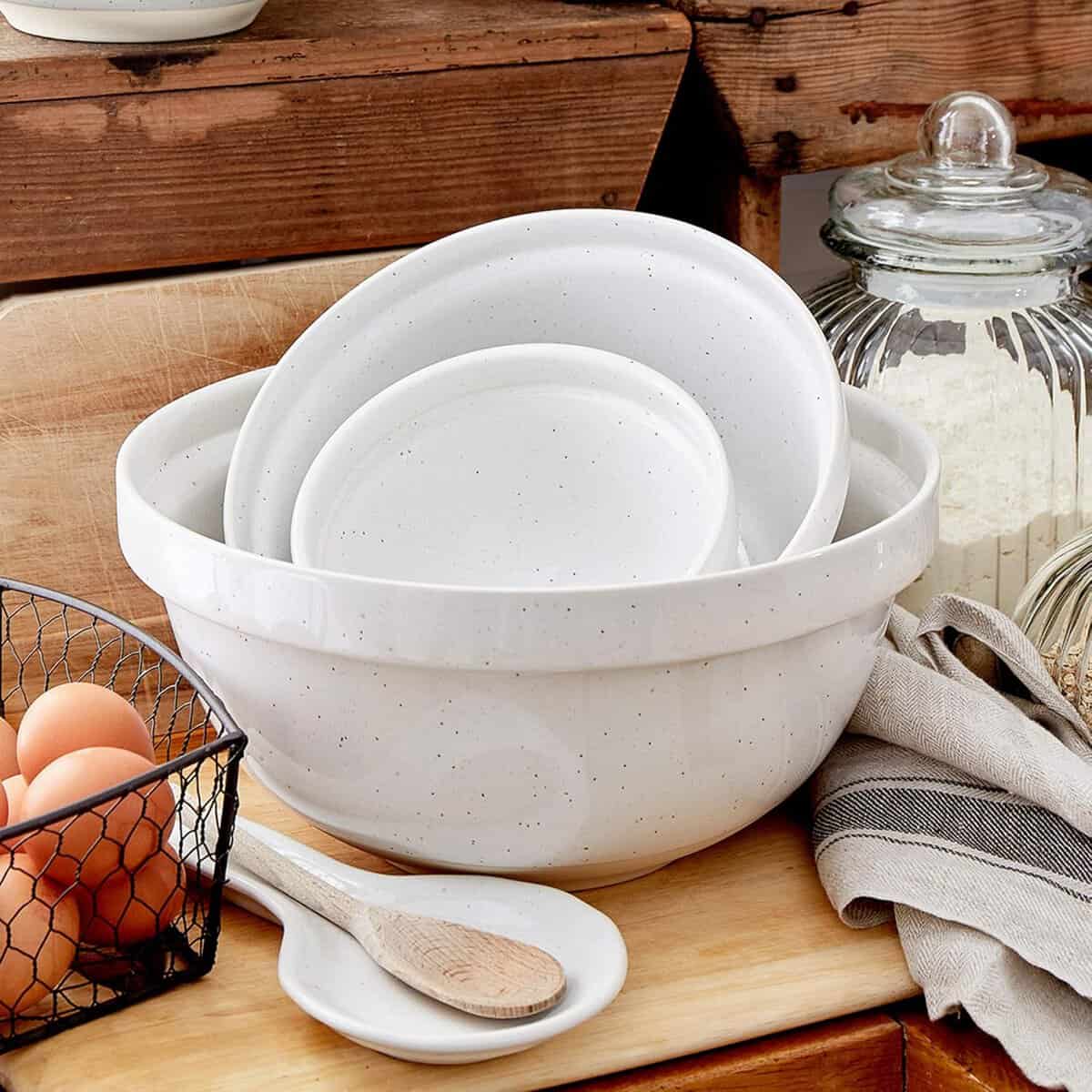 A set of white ceramic bowls and plates resting inside a larger bowl on a wooden counter. Nearby, a wire basket holds brown eggs, and a glass jar filled with flour sits next to a striped cloth and a wooden spoon.