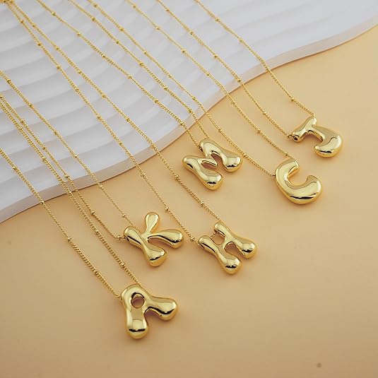 Gold necklaces with pendants shaped like letters R, K, M, H, C, and J are displayed on a soft beige surface with a white textured background.