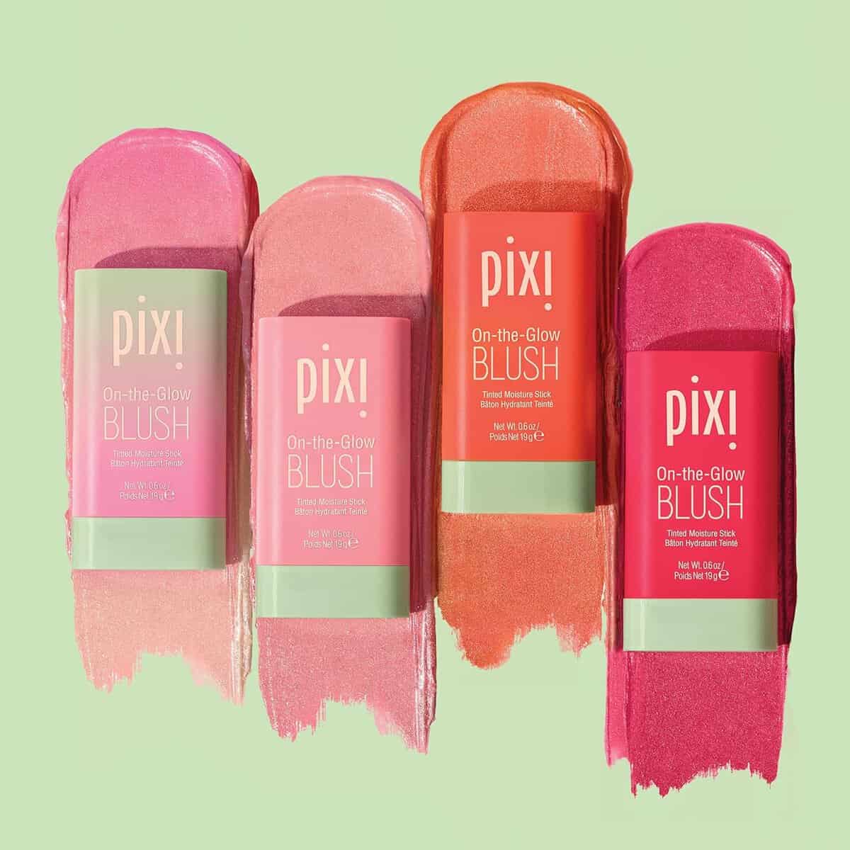 Four Pixi On-the-Glow Blush tubes are displayed against a green background. Each stick is shown with a swatch of its color: light pink, pink, coral, and rosy pink, highlighting a creamy texture.