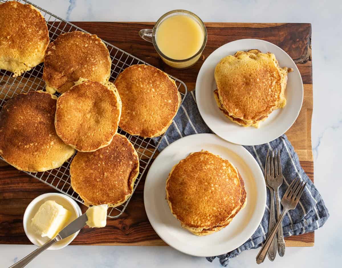 A wooden table with a wire rack holding several golden-brown pancakes. Two plates, each with a stack of pancakes, are nearby. Forks and knives are placed on a blue cloth, with a cup of orange juice and a small dish of butter also visible.