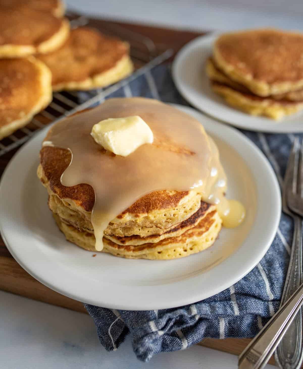 A stack of pancakes on a white plate, topped with melted butter and syrup. More pancakes are in the background on a wire rack and a small plate. A blue and white cloth and silverware are placed nearby.