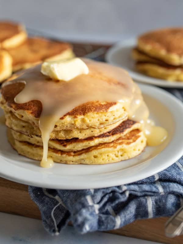 A stack of cornmeal pancakes topped with melting butter and syrup on a white plate. There are more pancakes in the background on a cooling rack and a separate plate. Forks are placed beside the dish on a striped cloth.