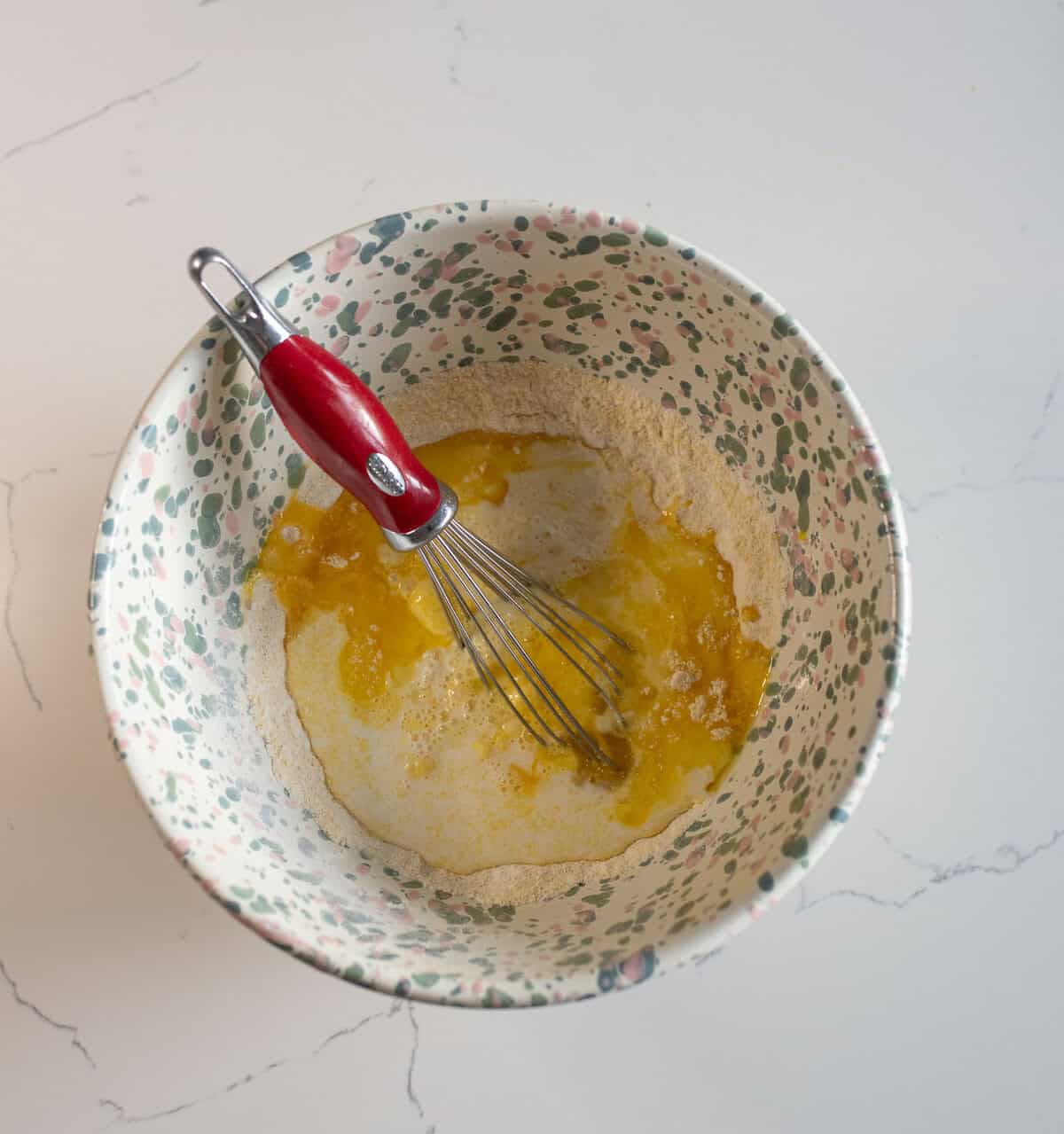 A colorful mixing bowl with a whisk and a mixture of eggs, flour, and liquid ingredients. The bowl has a floral pattern, and the whisk has a red handle. The background is a white, lightly marbled countertop.