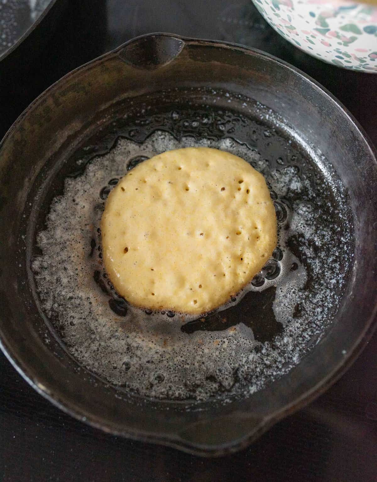 A pancake batter cooking in a black skillet with melted butter bubbling around the edges. The pancake is light brown and starting to form small bubbles on its surface.