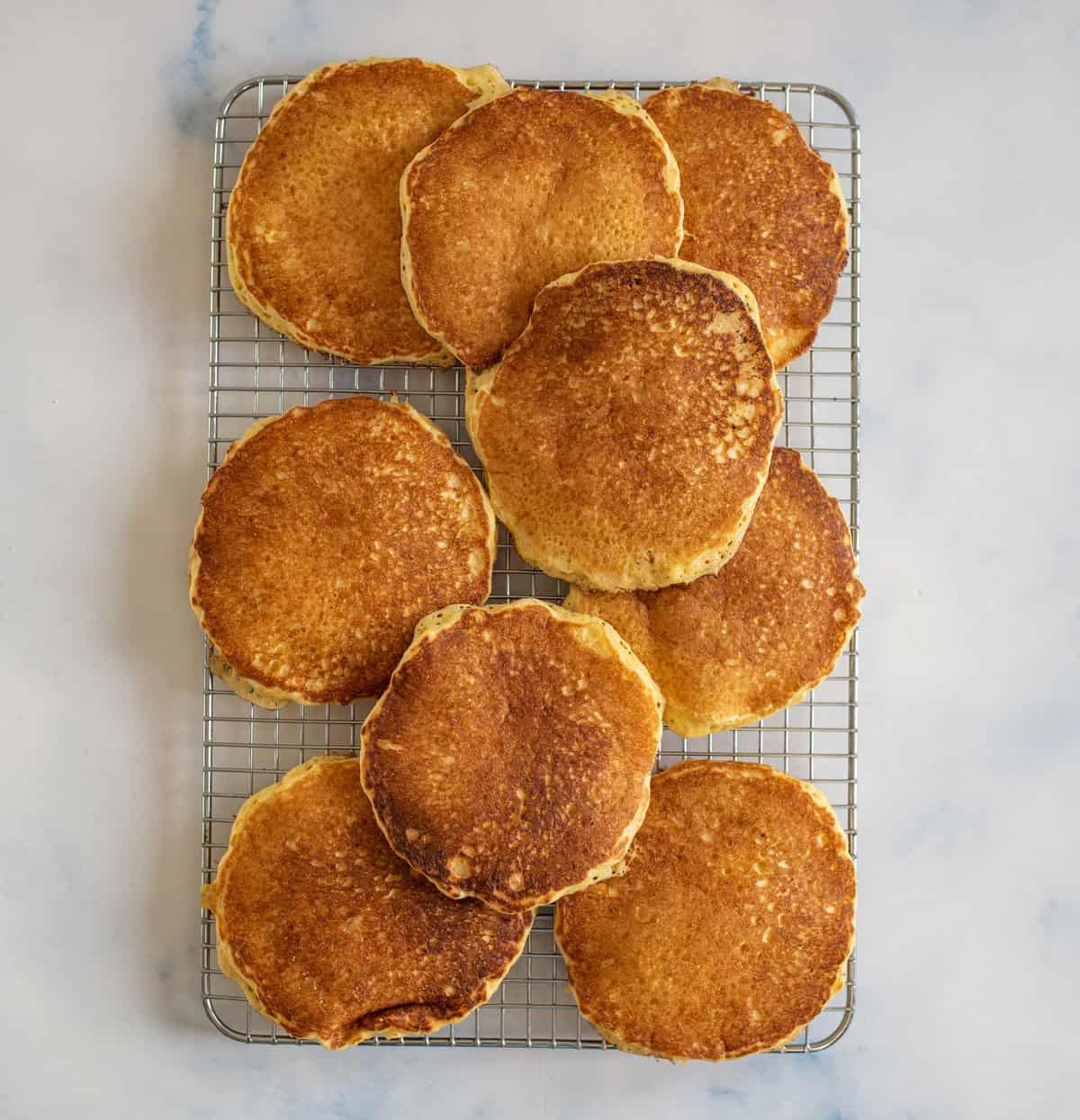 A cooling rack with nine golden-brown pancakes stacked neatly on a light marble surface. The pancakes have a slightly crisp texture and are evenly cooked, showcasing a homemade appearance.