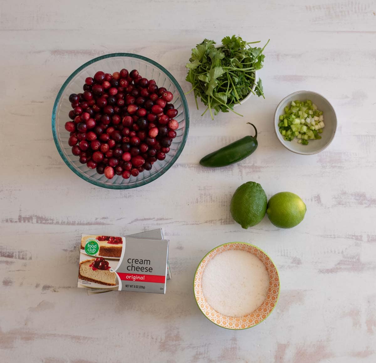 A bowl of cranberries, fresh cilantro, a jalapeño, and chopped green onions are on a white surface. Nearby are a pack of cream cheese, a lime, a lemon, and a small bowl of sugar.