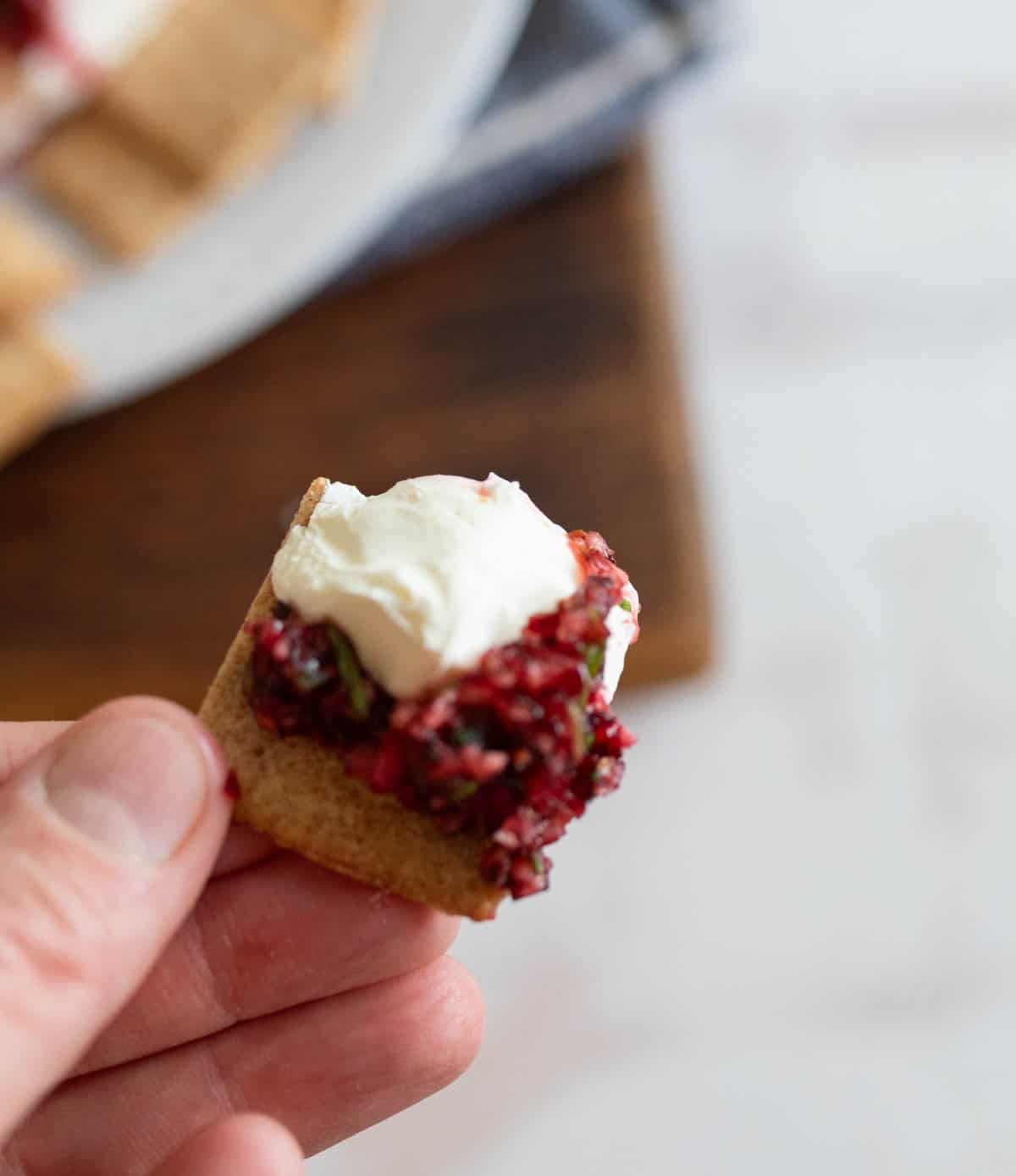 A hand holds a cracker topped with raspberry jam and a dollop of cream cheese. The background shows a blurred wooden surface with more crackers.