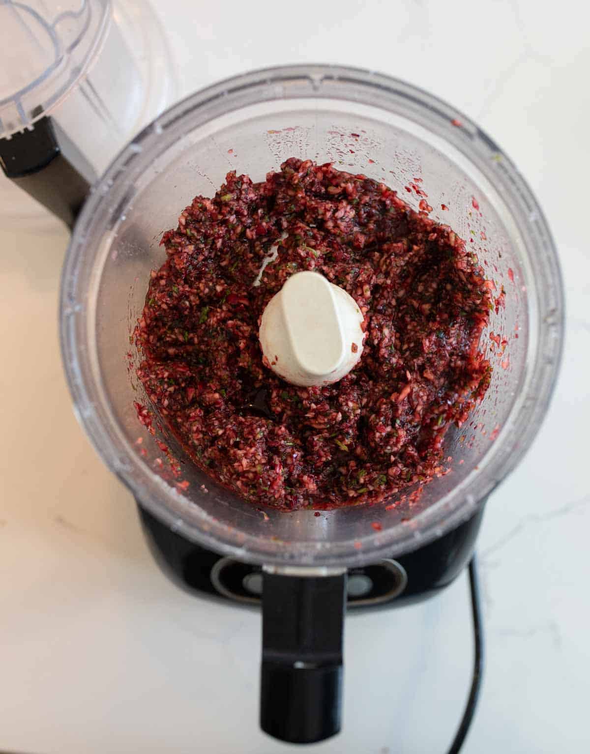 A top view of a food processor containing a finely chopped mixture of red and green ingredients, likely a relish or salsa, on a white countertop.