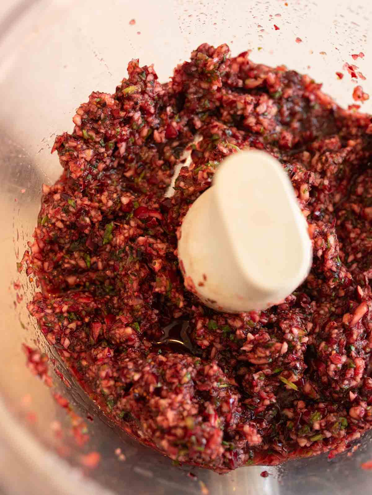 Close-up of a food processor containing a finely chopped blend of cranberries, nuts, and herbs, resulting in a textured mix in shades of red, green, and brown.