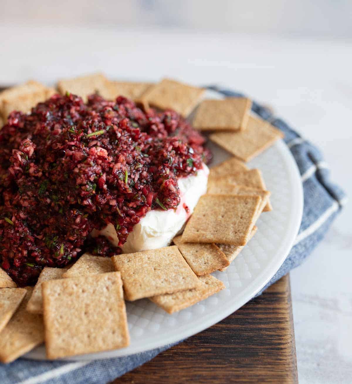 A plate of crackers is arranged around a mound of creamy dip topped with a reddish, finely chopped mixture with herbs, on a white and blue background.