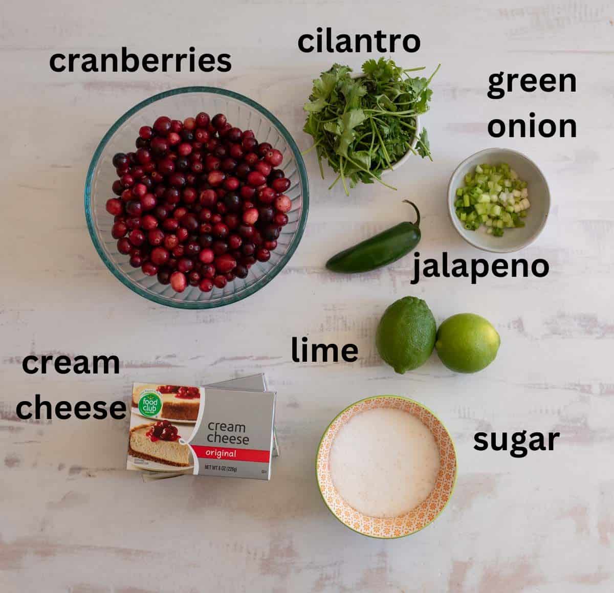 A top view of ingredients on a table: a bowl of cranberries, cilantro bunch, chopped green onion, a jalapeño, two limes, cream cheese package, and a small dish of sugar. Each item is labeled in text.