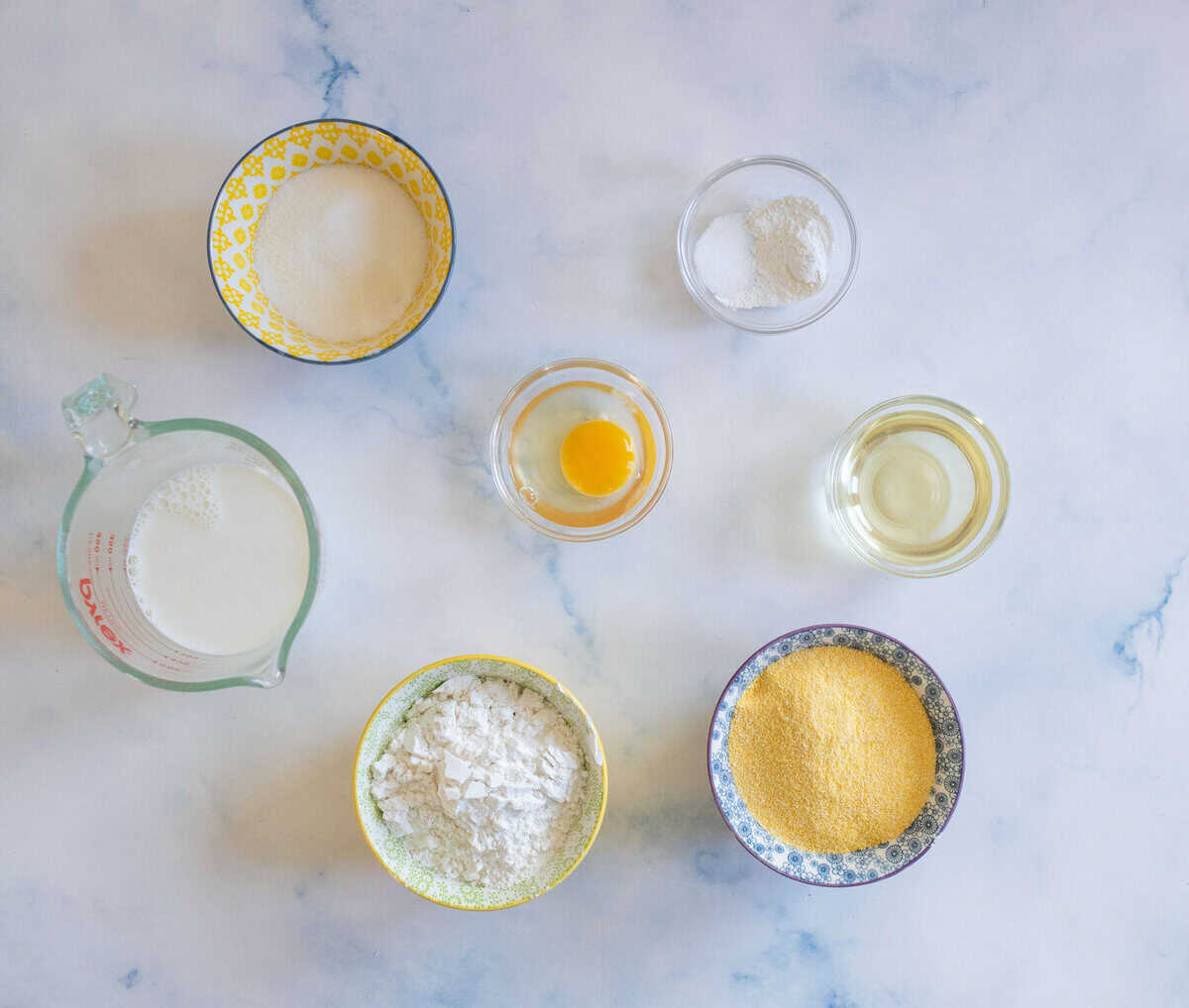 Top-down view of baking ingredients on a light surface: a bowl of sugar, a small bowl of salt, a glass of oil, a cup of milk, an egg yolk in a bowl, a bowl of flour, and a bowl of cornmeal.