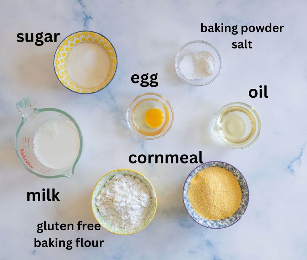 Top-down view of ingredients for a recipe on a marble surface: sugar, gluten-free baking flour, cornmeal, milk, egg, oil, baking powder, and salt, each labeled with text near their respective bowls.