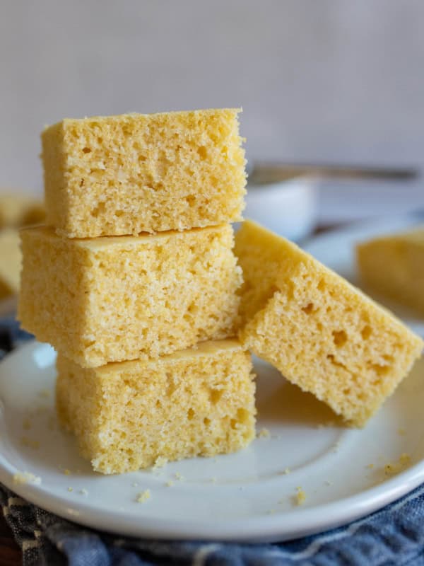 A stack of four square pieces of gluten free cornbread sits on a white plate. The cornbread has a light, fluffy texture and a golden yellow color. A fork and more pieces of cornbread are visible in the blurred background.