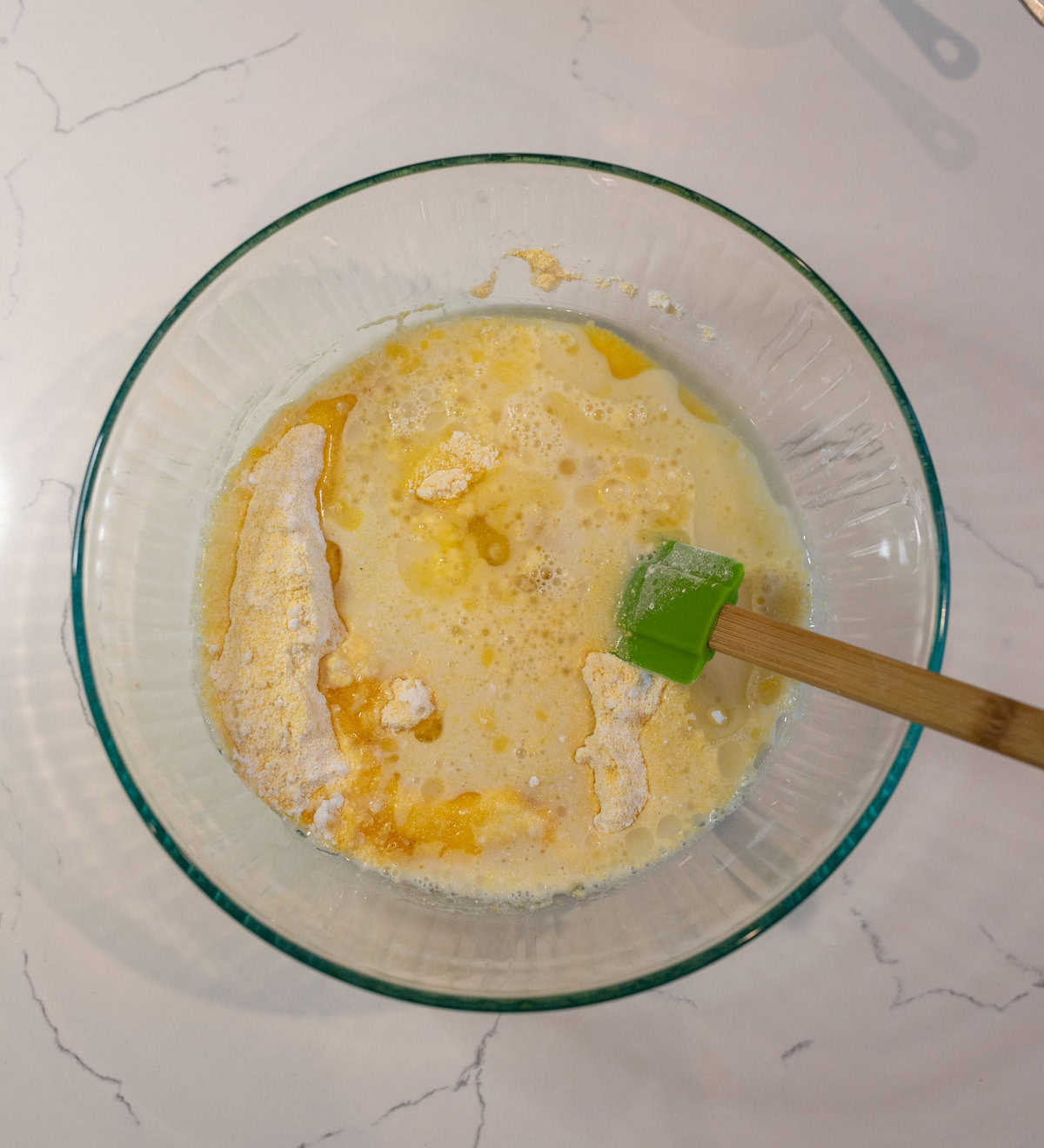 A glass bowl containing a mixture of flour, eggs, and liquid ingredients sits on a marble countertop. A green spatula with a wooden handle is resting in the bowl, suggesting the preparation of a batter.