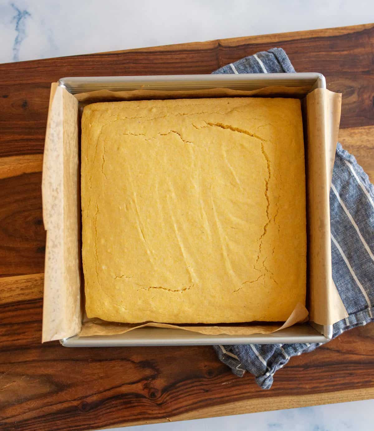 A freshly baked, square cornbread in a parchment-lined baking pan sits on a wooden cutting board. A blue-striped cloth is partially visible underneath the pan. The cornbreads surface is golden and slightly cracked.