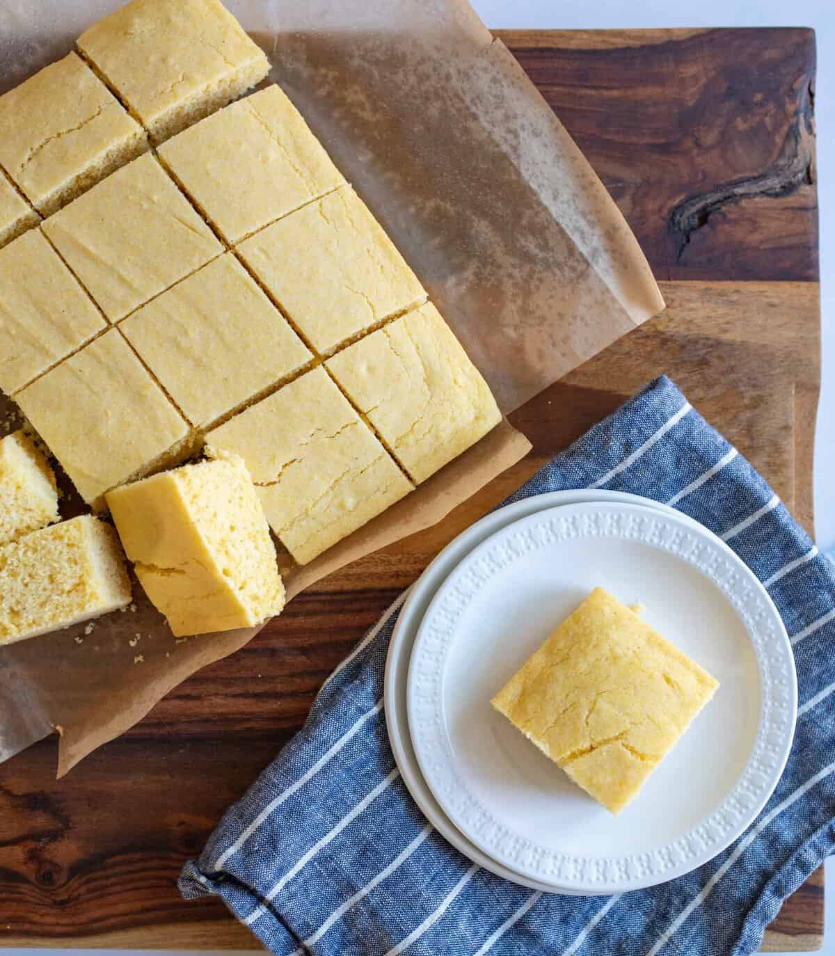 Sliced cornbread on parchment paper atop a wooden board. A single piece is placed on a white plate resting on a blue and white striped cloth.