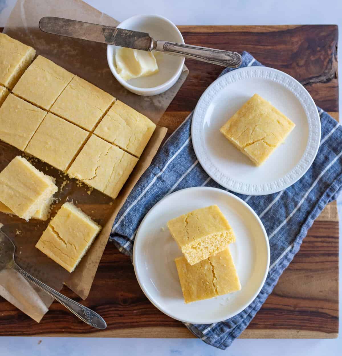 Sliced cornbread on a wooden board with two plates of cornbread squares beside it. A butter dish with a knife rests nearby on a blue cloth.