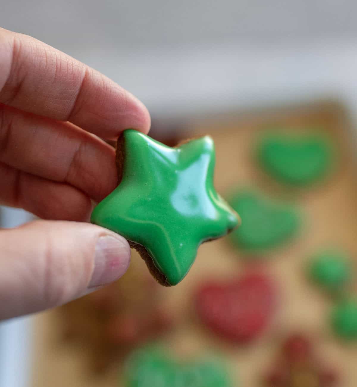 A hand holding a star-shaped cookie with glossy green icing. Blurred in the background are more decorated cookies in various shapes and colors on a tray.