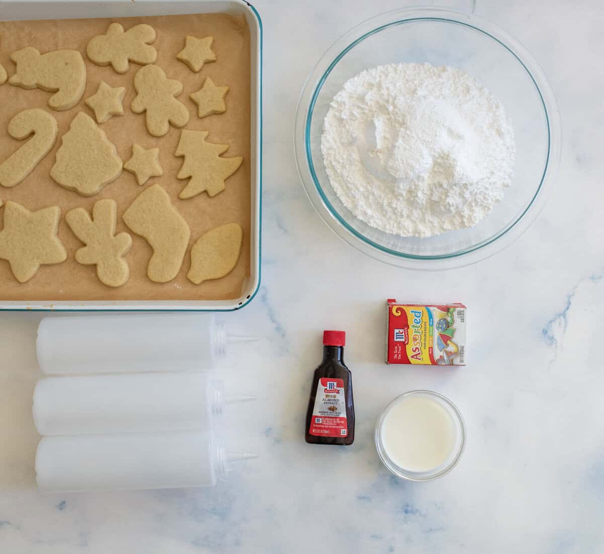 Overhead view of a baking setup showing a tray of baked cookies in various shapes, a bowl of powdered sugar, three squeeze bottles, a bottle of vanilla extract, a packet of food coloring, and a small bowl of milk on a marble surface.