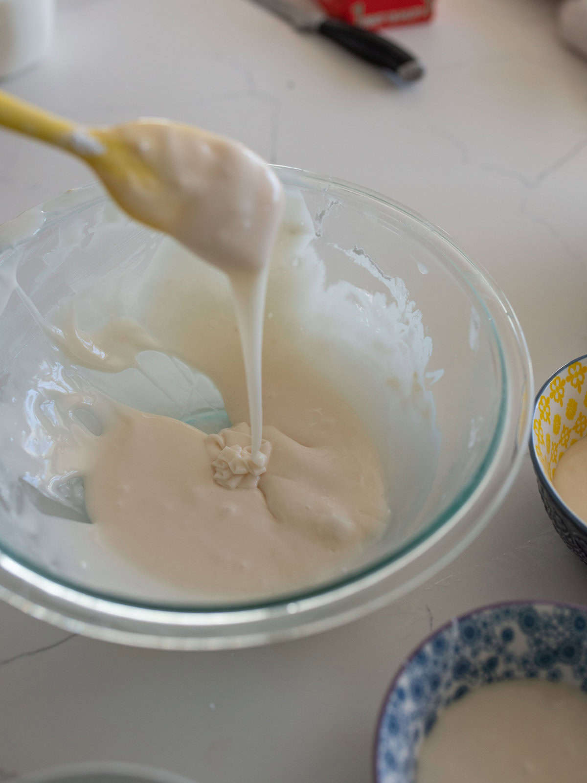 A glass bowl with thick, creamy white batter is being stirred with a yellow spatula. More batter drips from the spatula back into the bowl. In the background, two small patterned bowls contain additional batter.