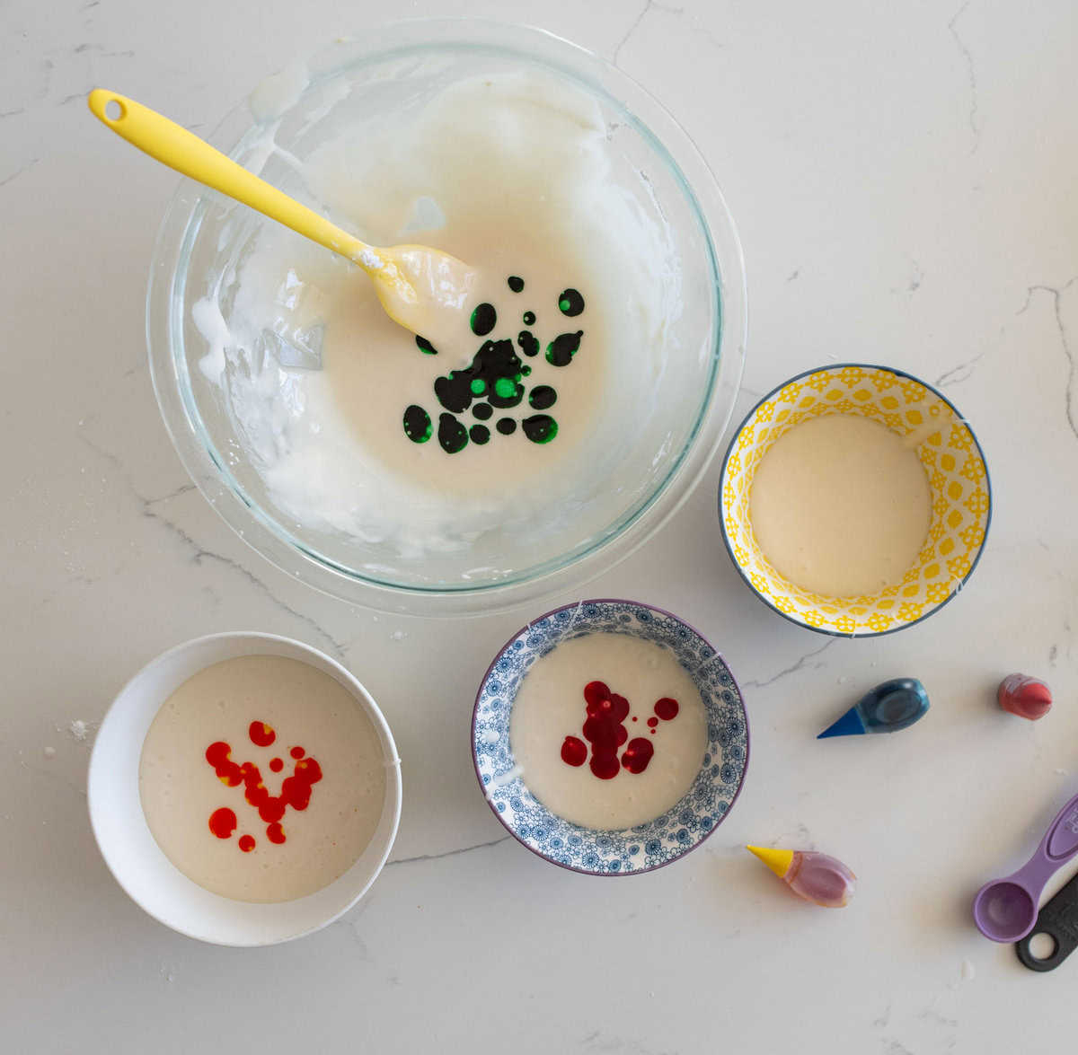 Four bowls with white batter, each with different food coloring: green, yellow, blue, and red. A yellow spoon in the largest bowl. Three food coloring bottles and measuring spoons nearby on a white countertop.