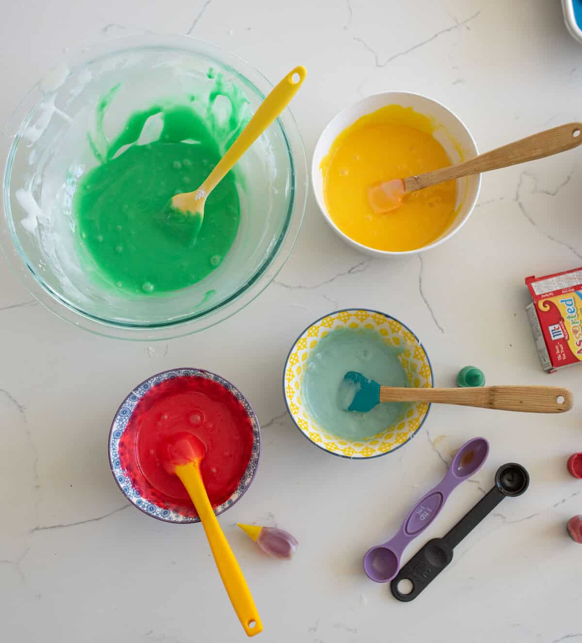 Four bowls with colorful cake batter—green, yellow, red, and light blue—each with a spatula. Measuring spoons and food coloring bottles are visible on the marble countertop.