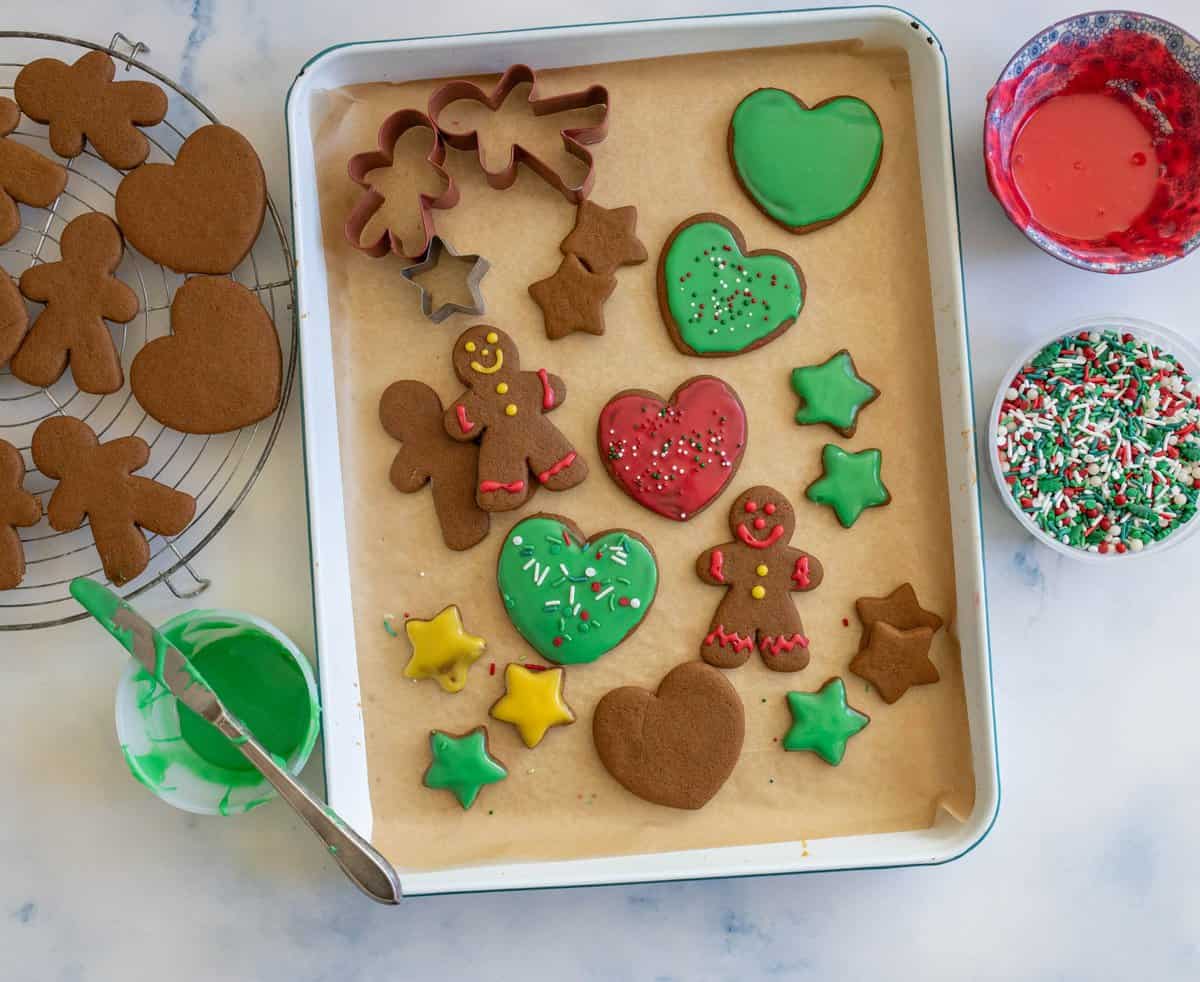 Baking tray with decorated gingerbread cookies in heart, star, and gingerbread person shapes. Icing is red, green, and white with sprinkles. Extra cookies on a cooling rack and bowls of icing and sprinkles are nearby.