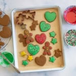 A baking tray with decorated gingerbread cookies in various shapes: hearts, stars, and gingerbread people. Green and red icing for gingerbread cookies with sprinkles. Nearby are bowls of red and green icing and a bowl of sprinkles.