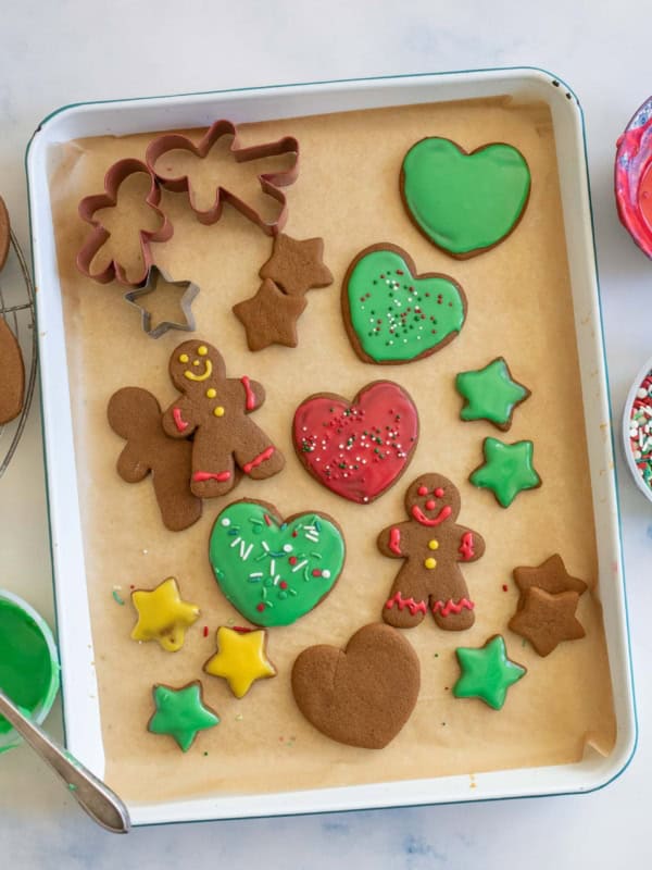 A baking tray with decorated gingerbread cookies in various shapes: hearts, stars, and gingerbread people. Green and red icing for gingerbread cookies with sprinkles. Nearby are bowls of red and green icing and a bowl of sprinkles.