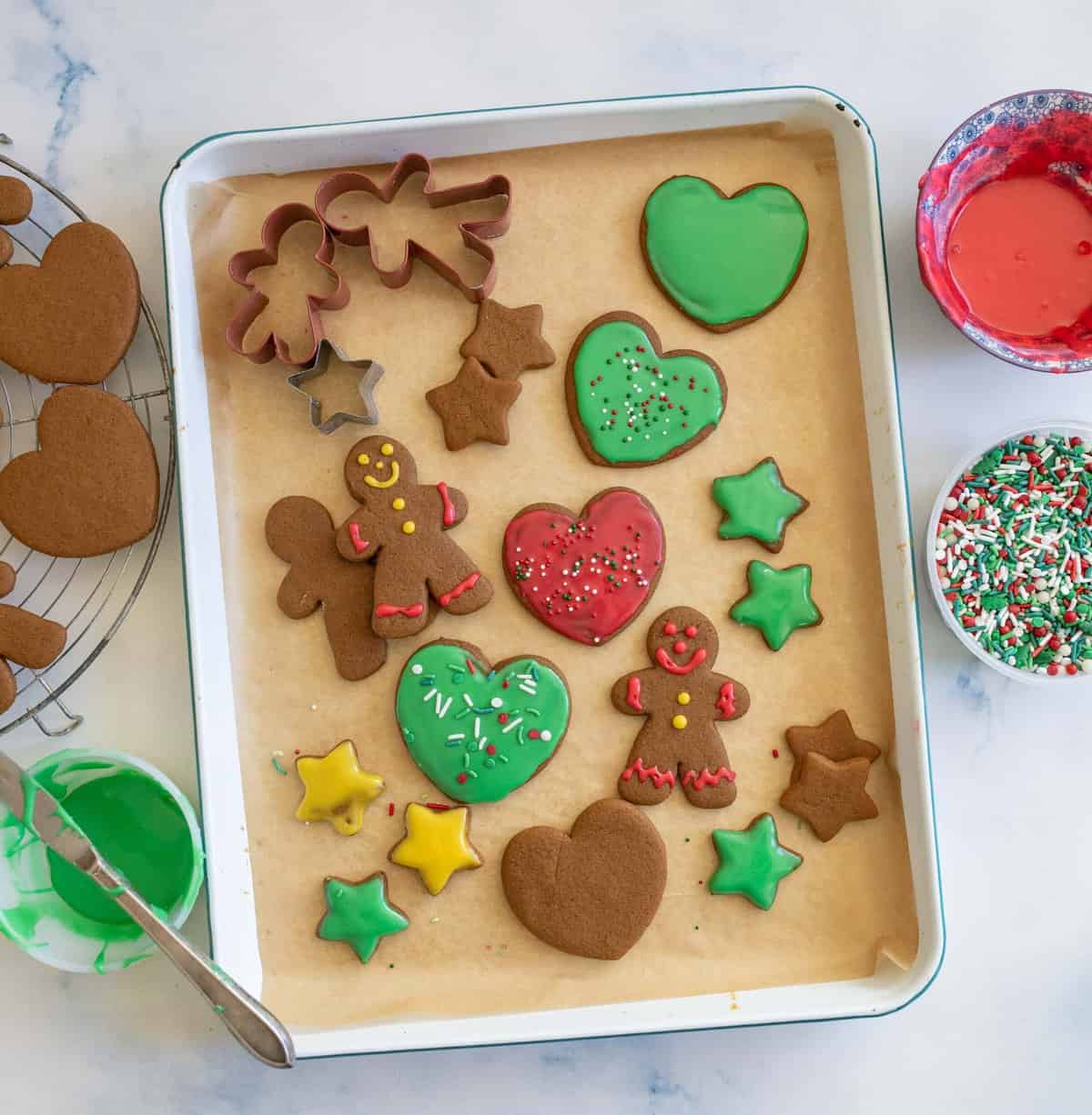 A baking tray with decorated gingerbread cookies in various shapes: hearts, stars, and gingerbread people. Green and red icing for gingerbread cookies with sprinkles. Nearby are bowls of red and green icing and a bowl of sprinkles.