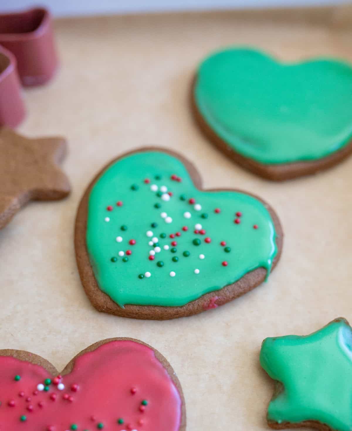 Heart-shaped cookies with green and red icing are decorated with colorful sprinkles. The cookies are on a parchment paper-lined surface, alongside cookie cutters.